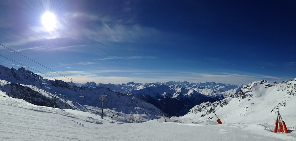 Panoramic sunny view from the top of the Mauriennaise red slope near Rosaël chairlift in Orelle, showing freshly groomed snow, snow cannons, and layered alpine peaks under a clear blue sky.