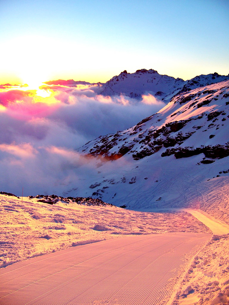 Golden sunset light and mountain shadows on the Mauriennaise ski run in Orelle after a day of skiing, with snowgroomer tracks above a sea of clouds.