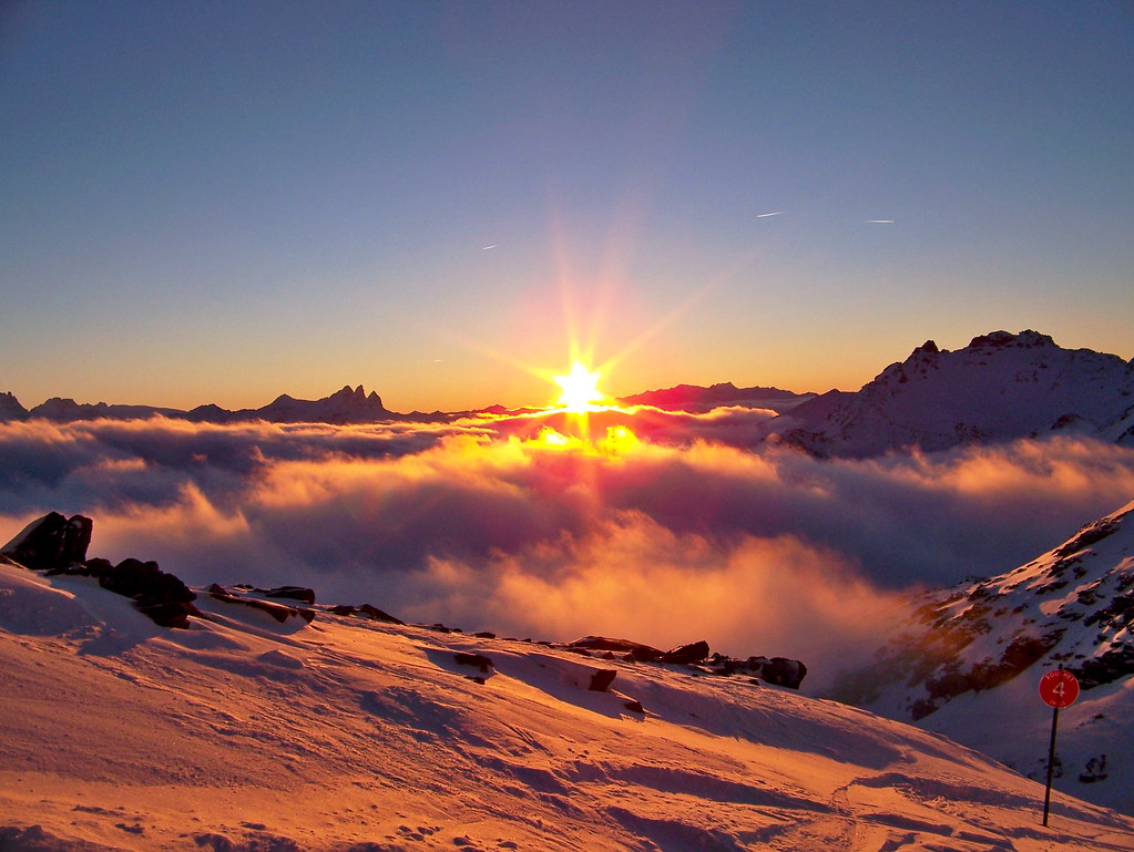 Orange sunset over a sea of clouds on the Bouchet red slope starting from Col du Bouchet in Orelle, with Cime Caron on the right and Aiguilles d'Arves on the horizon.