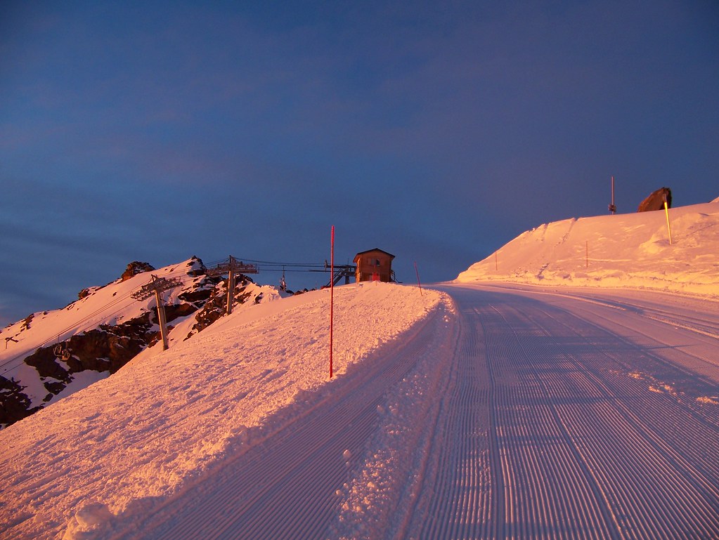 Golden hour view at Col du Bouchet in Orelle at 3230m altitude, showing the top station of the Bouchet chairlift, freshly groomed red slope, and the start of the zipline.