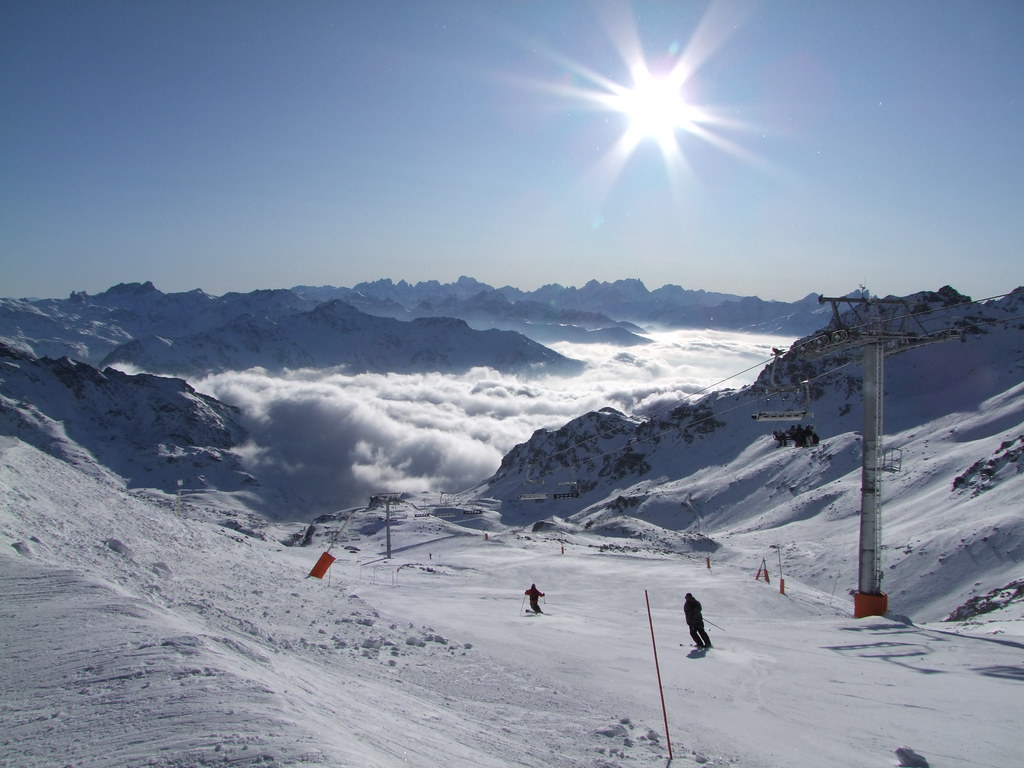 Two skiers carving down the Mauriennaise red slope viewed from Gentianes run in Orelle, with Col Rosaël chairlift, sunny blue sky, and a sea of clouds covering the valley below.