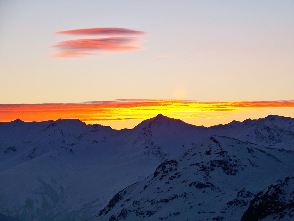 High-altitude twilight panorama in Orelle, with night falling over dark mountain silhouettes and soft orange clouds reflecting the set sun over distant valleys.