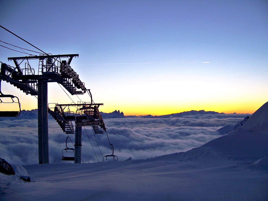 Dusk view from Col Rosaël in Orelle, showing the chairlift emerging from the steep slope alongside a sea of clouds and mountain silhouettes at sunset.