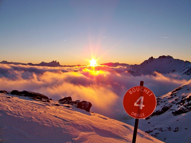 Red slope marker board number 4 on the Bouchet run in Orelle at sunset, with a sea of clouds and the Aiguilles d'Arves in the background.