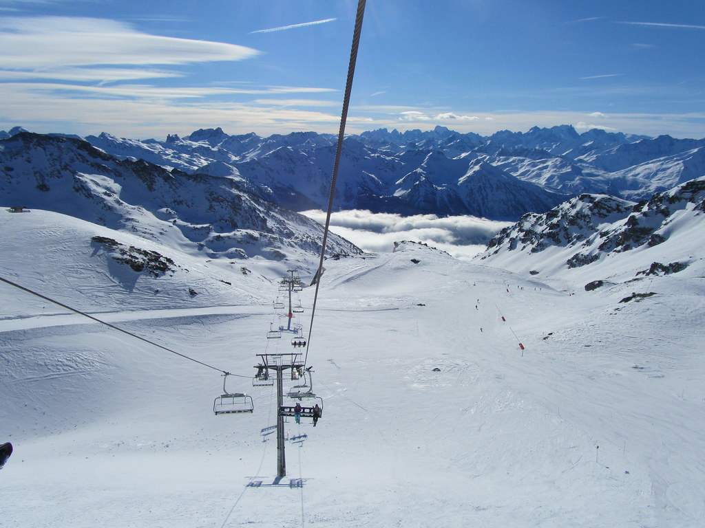 Onboard view from the Col de Rosaël chairlift seat in Orelle, showing uncrowded ski slopes, jagged mountain horizons, and a bright blue sky.
