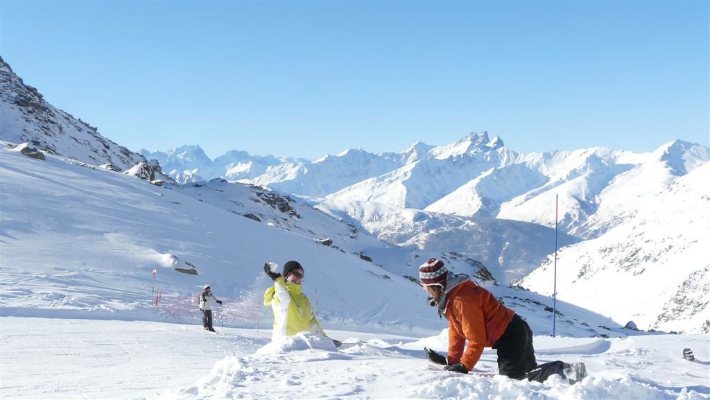 Two friends having a fun snowball fight on the side of the Peyron ski slope in Orelle, with clear blue skies and the Aiguilles d'Arves on the horizon.
