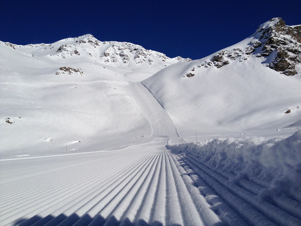 Ground-level closeup of freshly groomed corduroy snow on a quiet morning in Orelle, with blue sky and rocky mountain peaks in the background.