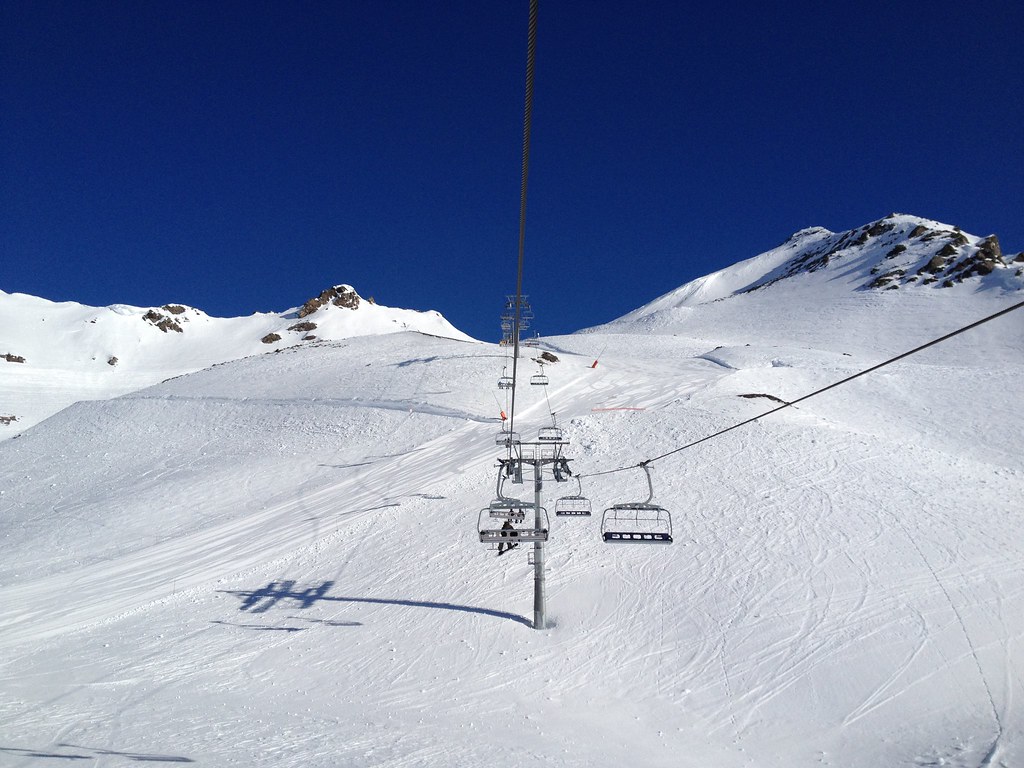 View from the Col de Rosaël chairlift in Orelle climbing over the Mauriennaise run towards the Val Thorens crossover, with a snowboarder ahead on a sunny day.