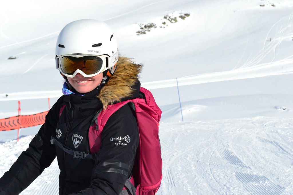 Front view portrait of a smiling female skier in Orelle gear standing on a sunlit slope under a clear blue sky.