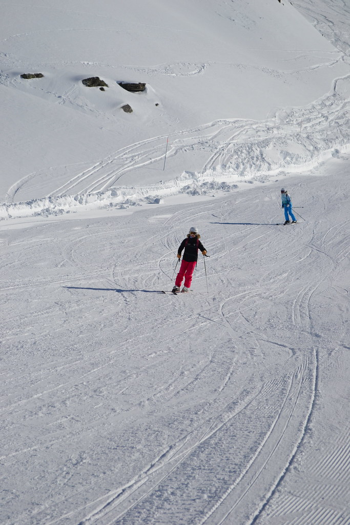 Distant view of two skiers descending a wide sunny slope in Orelle with snow-capped alpine mountains in the background.