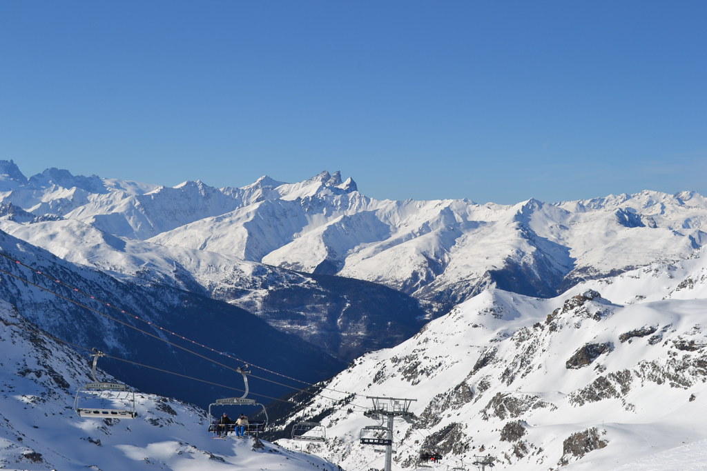 View of the Aiguilles d'Arves peaks above Valloire seen from Orelle ski slopes, with skiers riding a chairlift under a clear blue sky.